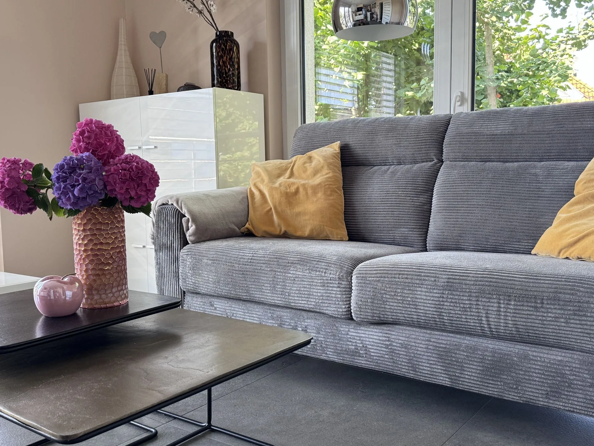 Helles Wohnzimmer der Guetsel Lodge mit grauem Sofa, warmen Kissen und frischen Hortensien